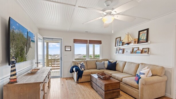 A living room with a beige sofa, wooden coffee table, and a television on a wooden cabinet. There is a ceiling fan, a view of the outdoors through large windows, and a shelf with framed pictures and a ship model. - Newly Renovated, 4 bed 2 bath, Pet Friendly Oceanfront w/ a Pool! (Brunswick)