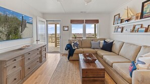 A spacious living room with a large window, a ceiling fan, and a wooden coffee table. - Newly Renovated, 4 bed 2 bath, Pet Friendly Oceanfront w/ a Pool! (Brunswick)