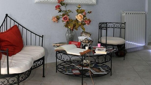 Un salón con un espejo de marco blanco, una mesa de centro redonda con revistas, un jarrón con flores y dos sillas metálicas con cojines.