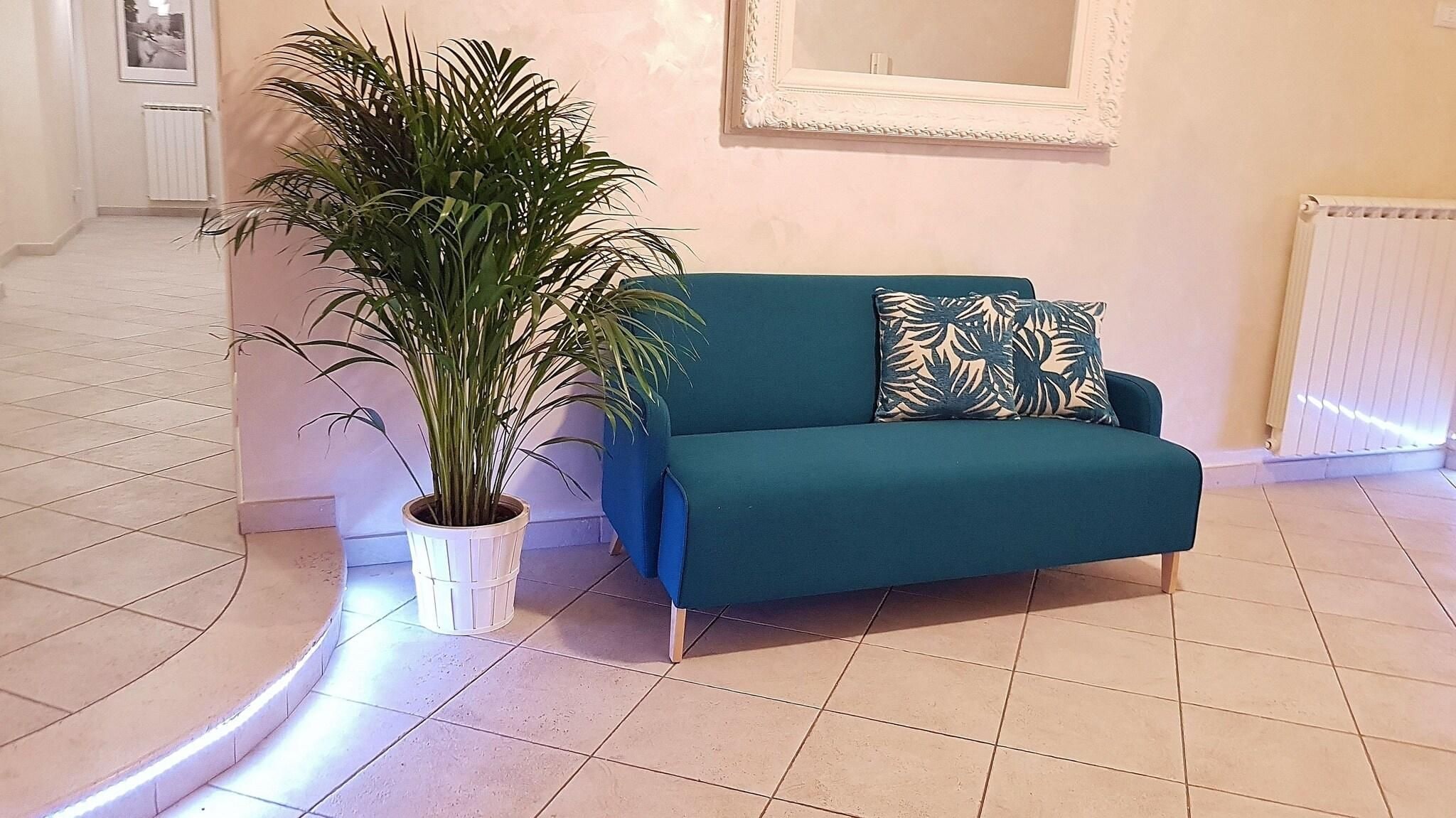 Un canapé de couleur turquoise agrémenté d’un coussin à motifs de feuilles, une plante en pot et un tableau encadré accroché au mur.