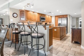 A modern kitchen with wooden cabinets, a stainless steel refrigerator, and a bar area with stools.