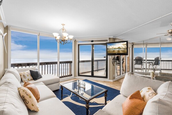 A spacious living room with a large sofa, a coffee table, and a balcony with a view of the ocean.