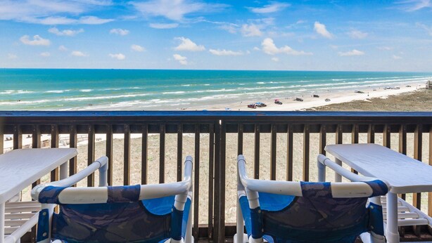 Une vue sur la plage depuis un balcon, avec deux chaises et une table, donnant sur une plage de sable où se trouvent quelques personnes et des véhicules.