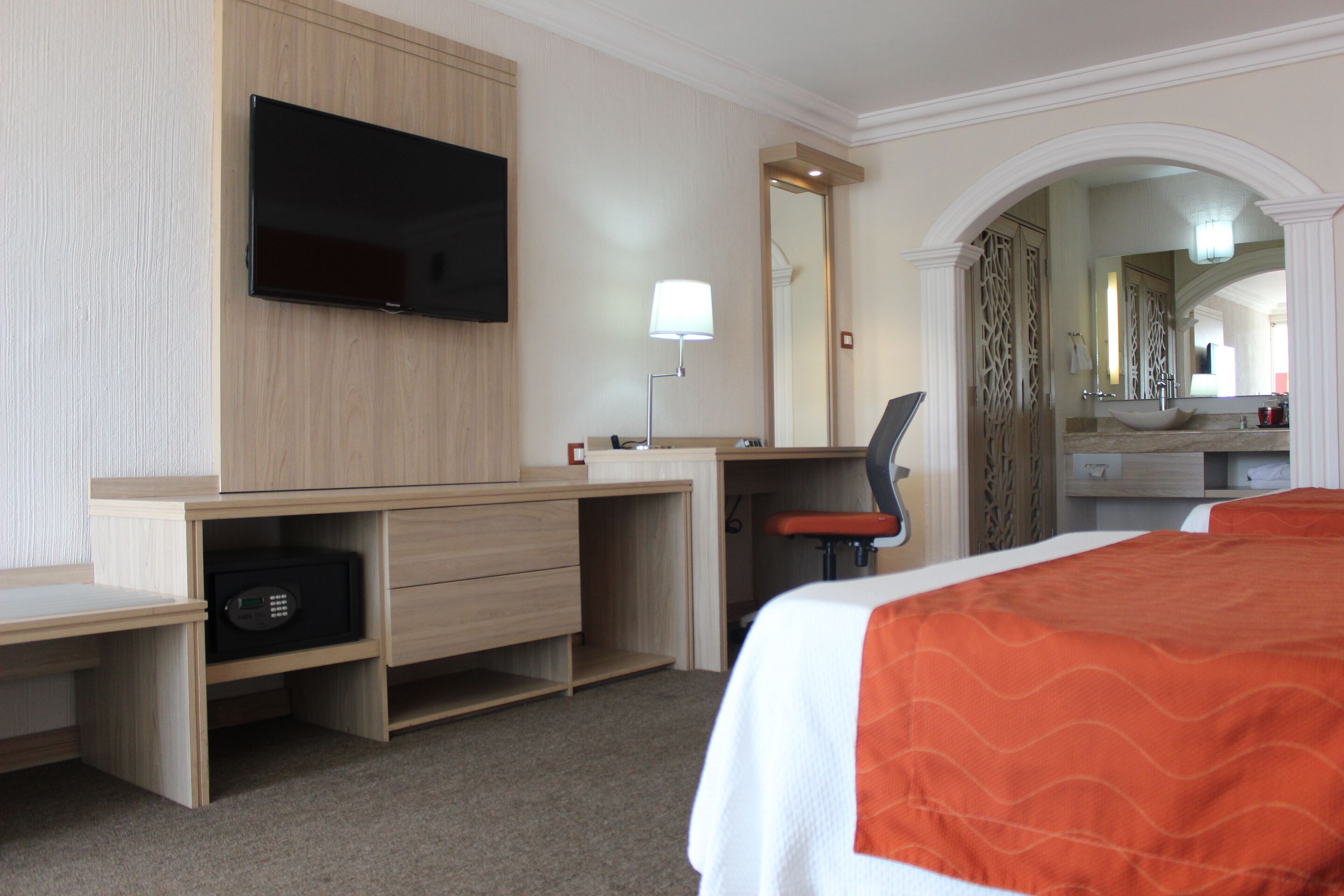 Una camera d'albergo con un letto, una scrivania con un televisore, una lampada, una sedia e un bagno visibile attraverso un arco.