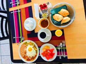 A table with a meal consisting of a bowl of enchiladas, a plate of fruit, a cup of coffee, a bowl of soup, and a plate of bread rolls.