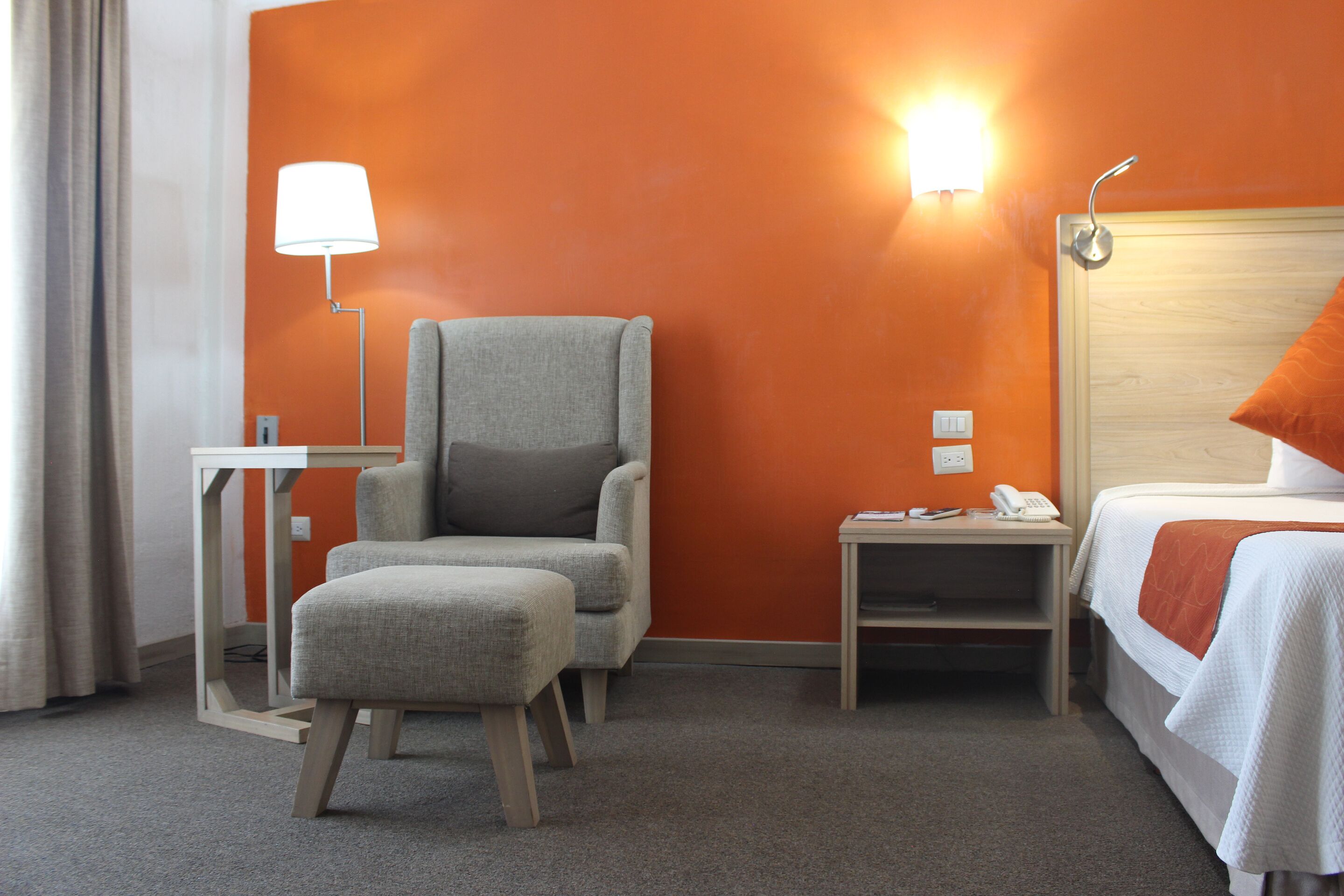 Une chambre d’hôtel avec un fauteuil gris, une petite table avec une lampe, une table de chevet avec un téléphone et un lit recouvert d’une couverture orange.