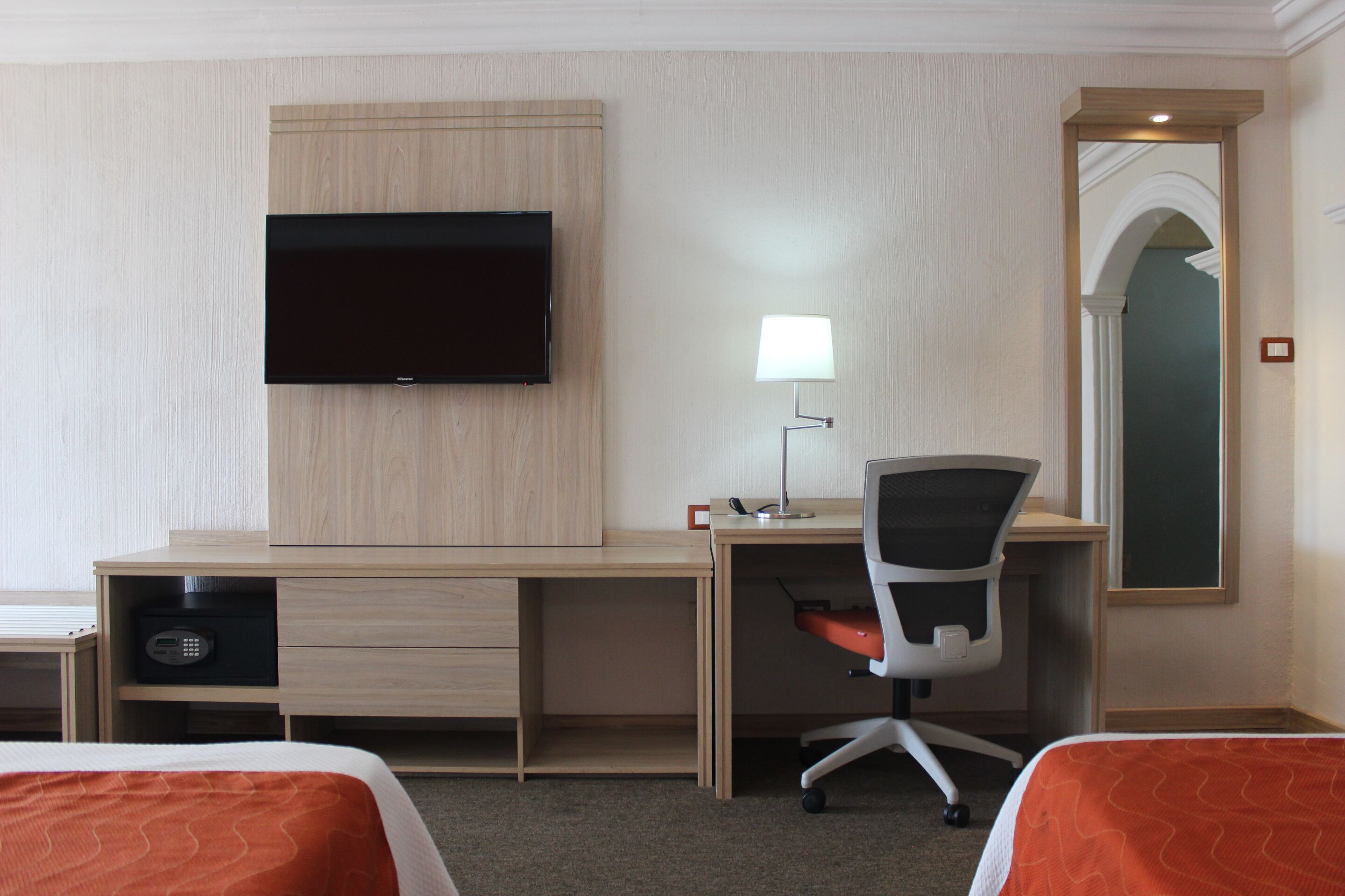 A hotel room with a wooden desk, a television, a desk lamp, a chair, and two beds with orange bedding.