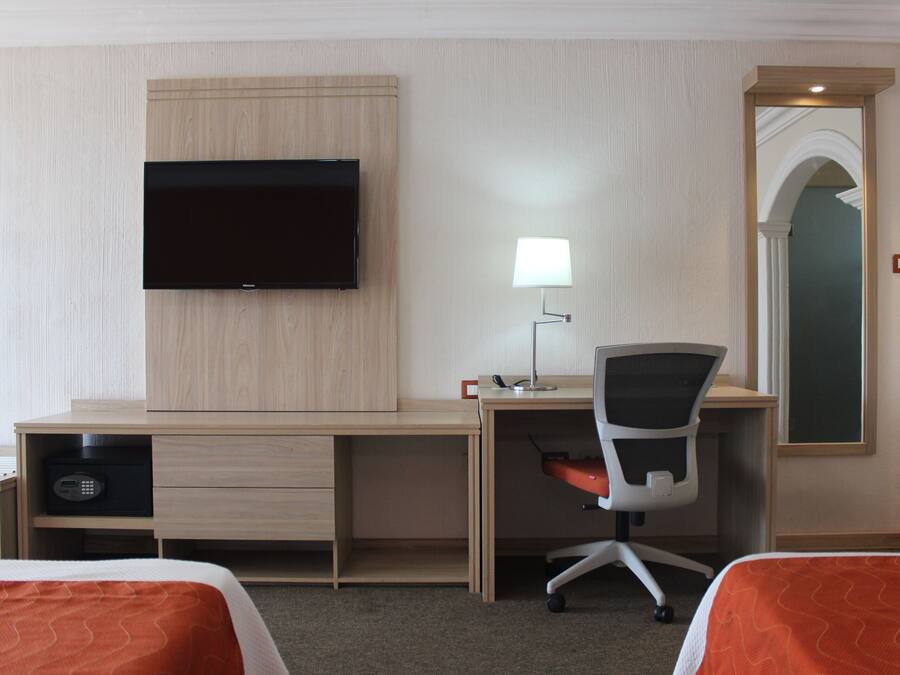 A hotel room with a wooden desk, a television, a desk lamp, a chair, and two beds with orange bedding.