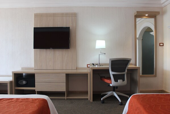 A hotel room with a wooden desk, a television, a desk lamp, a chair, and two beds with orange bedding.