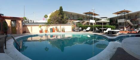 Una piscina con sillas de descanso y sombrillas, rodeada de edificios con balcones.