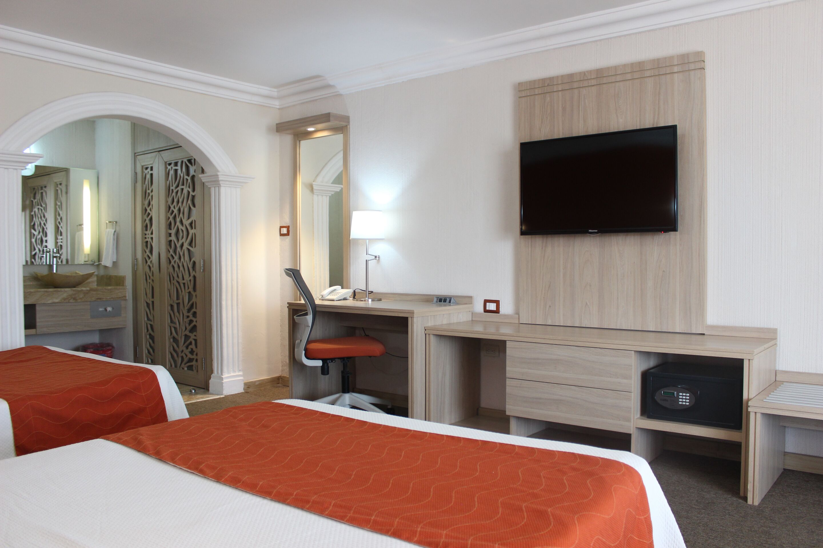 A hotel room with two beds, a desk, a chair, a television, a mirror, and a bathroom with a shower and sink.
