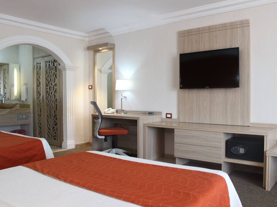 Habitación de hotel con dos camas, un escritorio, una silla, un televisor, un espejo y un baño con ducha y lavamanos.