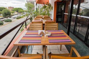 Un espacio de comedor al aire libre con mesas y sillas de madera, individuales a rayas y vistas a la calle y edificios al fondo.