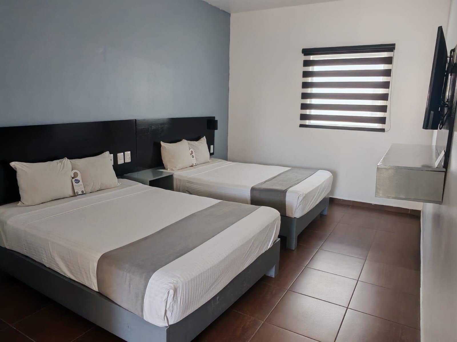 Habitación de hotel con dos camas, una mesita de noche, un televisor y una ventana con persianas.