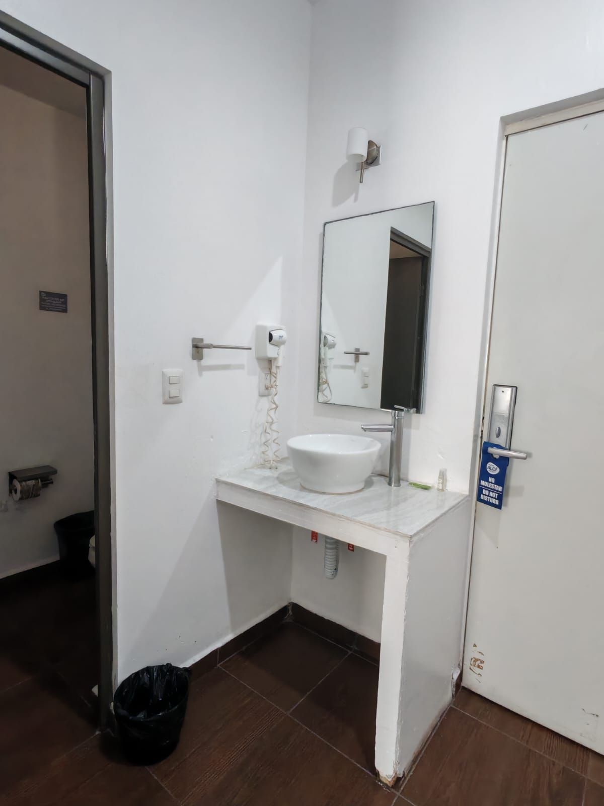 Un baño con lavamanos blanco, espejo y puerta con un cartel.