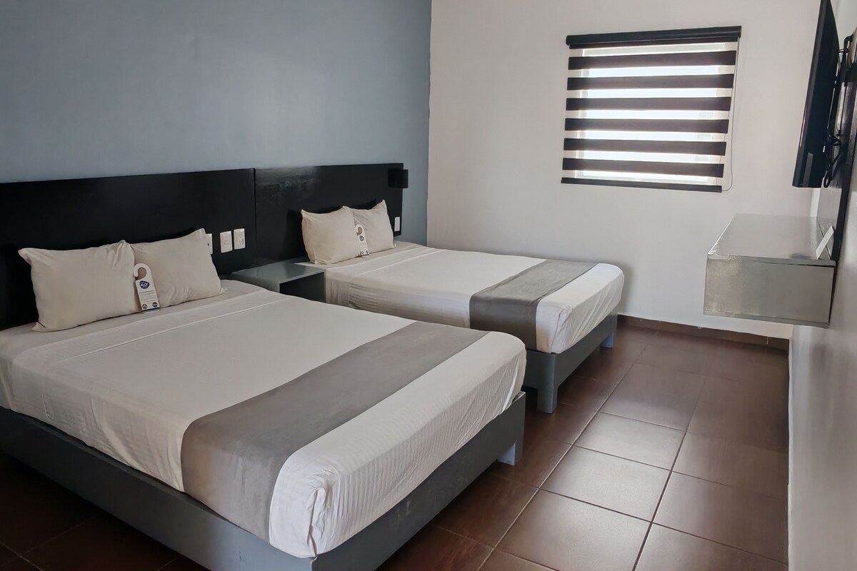 A hotel room with two beds, a nightstand, a wall-mounted TV, and a window with blinds.