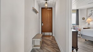 Ein schmaler Flur mit einer Holztür, einer Bank und einem Bett im anliegenden Zimmer.
