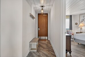 A narrow hallway with a wooden door, a bench, and a bed in the adjacent room.