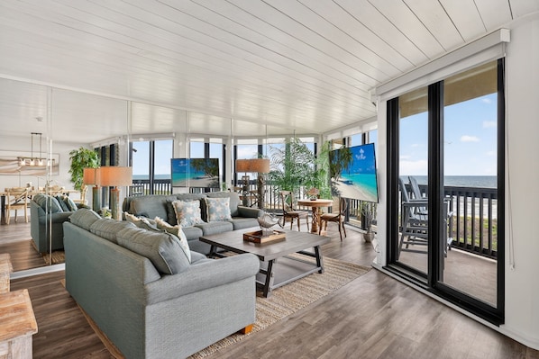 A spacious living area with a large sofa, coffee table, and dining table, featuring a view of the ocean through large sliding glass doors.