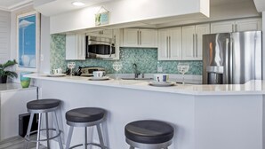 A modern kitchen with white cabinets, a stainless steel refrigerator, a microwave, and a tiled backsplash. There are three bar stools and a hanging light fixture.