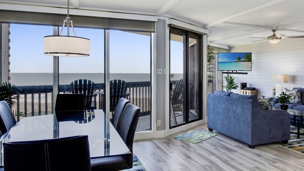 A modern living room with a glass dining table, a blue sofa, and a large window offering a view of the beach.