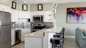 A modern kitchen with white cabinets, a stainless steel refrigerator, a microwave, and a gas stove. There is a bar area with stools and a colorful abstract painting on the wall. - Sandpiper Condominiums 1208, Panoramic Gulf Views w/ direct access to Mustang Island/Port A beaches. (Port Aransas)
