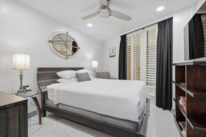 A bedroom with a bed, bedside lamps, a round wall decoration, a ceiling fan, and a window with blinds.