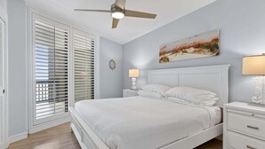 A bedroom with a bed, nightstands, a ceiling fan, a window with blinds, and a wall-mounted lamp.