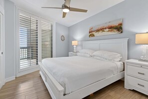 A bedroom with a bed, nightstands, a ceiling fan, a window with blinds, and a wall-mounted lamp. - Sandpiper Condominiums 1105, Panoramic Gulf Views w/ direct access to Mustang Island/Port A beaches. (Port Aransas)