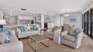 A modern living room with a sofa, armchair, and coffee table, a kitchen with white cabinets, and a dining area with a table and chairs.