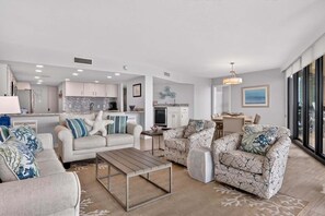 A modern living room with a sofa, armchair, and coffee table, a kitchen with white cabinets, and a dining area with a table and chairs. - Sandpiper Condominiums 1105, Panoramic Gulf Views w/ direct access to Mustang Island/Port A beaches. (Port Aransas)