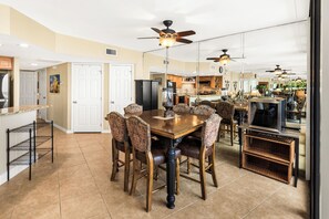 A dining area with a wooden table and patterned chairs, a ceiling fan, and a kitchen in the background.