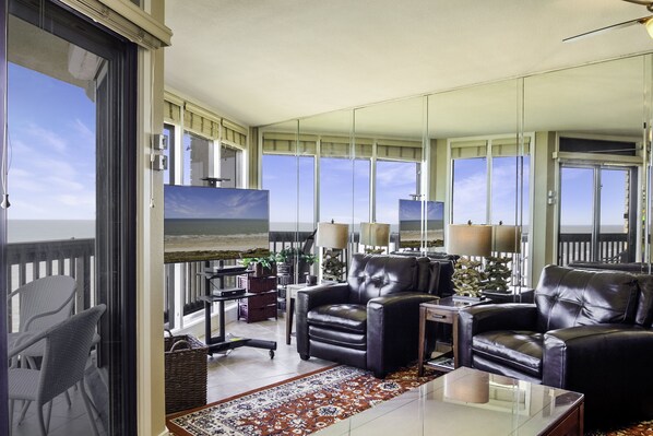Een ruime woonkamer met een groot raam dat uitkijkt op een balkon, leren banken, een bijzettafeltje en uitzicht op de oceaan.