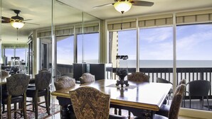 A dining area with a large table and chairs, large windows with a view, and ceiling fans. - Sandpiper Condominiums 1104L, Panoramic Gulf View w/ direct access to Mustang Island/Port A beaches. (Port Aransas)