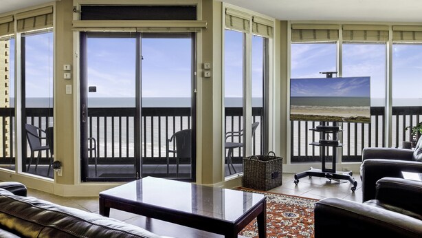 A corner room with a sliding glass door leading to a balcony, a flat-screen TV, a leather sofa, and a small table. - Sandpiper Condominiums 1104L, Panoramic Gulf View w/ direct access to Mustang Island/Port A beaches. (Port Aransas)