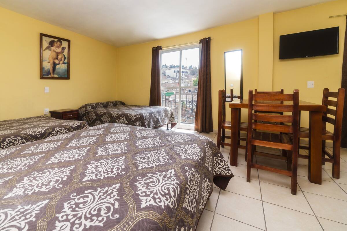 Habitación de hotel con dos camas, una mesa de comedor con sillas, un televisor en la pared y un balcón con vista a la ciudad.