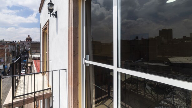 Un edificio con grandes ventanales que reflejan el cielo y las nubes, un balcón con barandilla de metal, y una vista a la calle con otros edificios y una estructura en forma de cúpula a lo lejos.