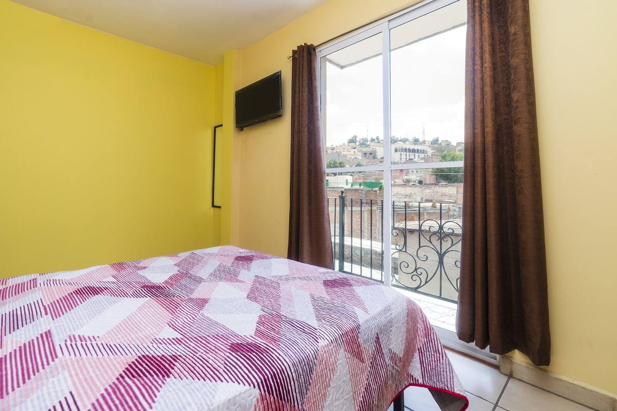 Una habitación con cama, televisor en la pared y balcón con cortinas.