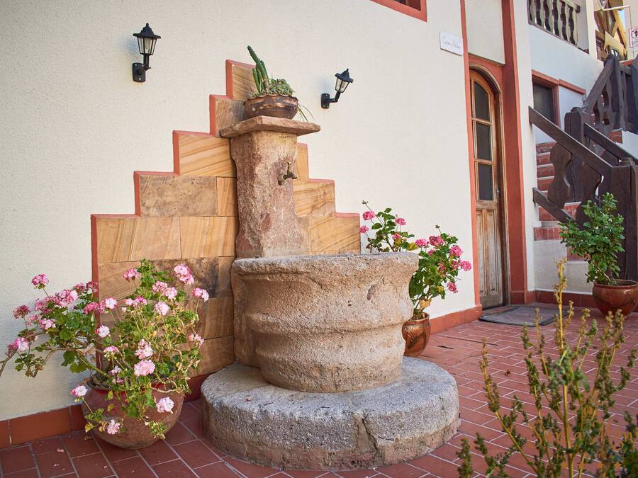 A stone fountain with a wooden staircase leading up to a door, surrounded by potted plants and flowers.