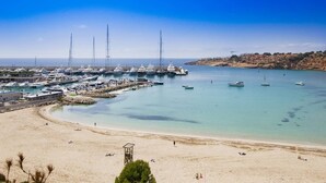 Een kustjachthaven met meerdere jachten afgemeerd, een zandstrand en een heldere blauwe lucht.