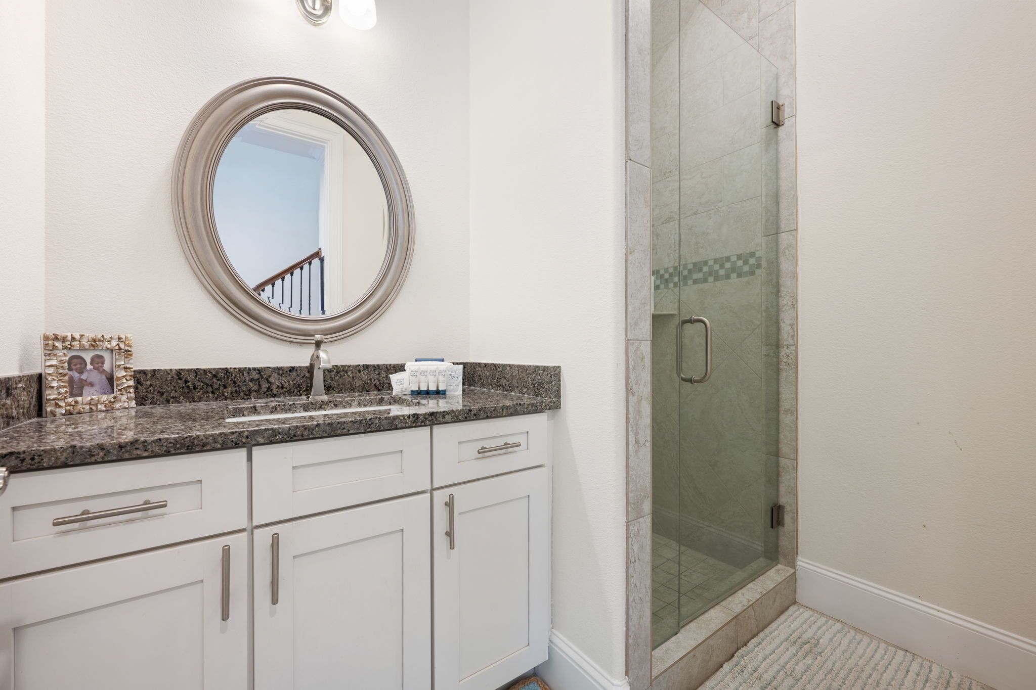 A bathroom with a granite countertop, a round mirror, a framed photo, and a glass shower enclosure.