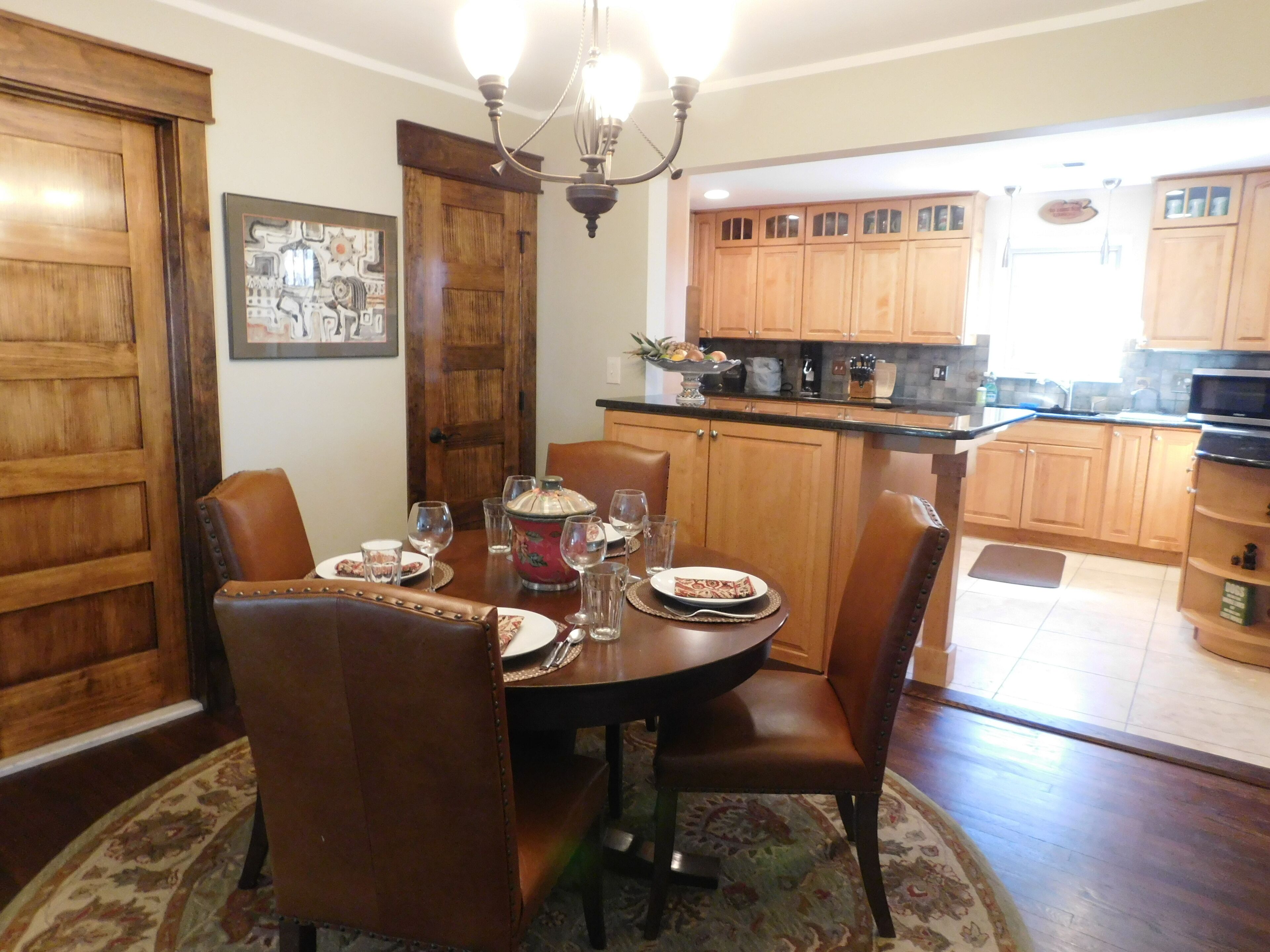 Un'area pranzo con un tavolo rotondo, sedie e un lampadario. Si nota una cucina sullo sfondo, con mobili a legno e un forno a microonde.