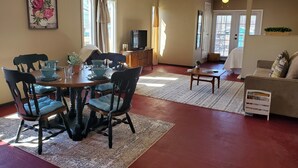A dining area with a wooden table, chairs, and a television. There is a sofa and a window with curtains in the living room.