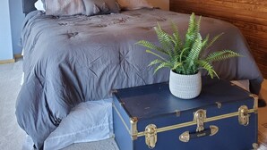 Un dormitorio con cama, cabecera, lámpara de pared, planta en maceta y maleta.