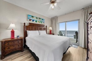 A bedroom with a large bed, wooden headboard, two bedside tables with lamps, a painting on the wall, and a balcony with a view.