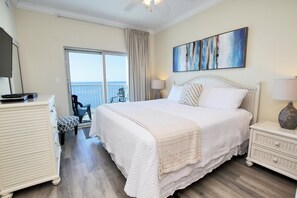 A bedroom with a large bed, a dresser, a nightstand, and a view of the sea through a sliding door.