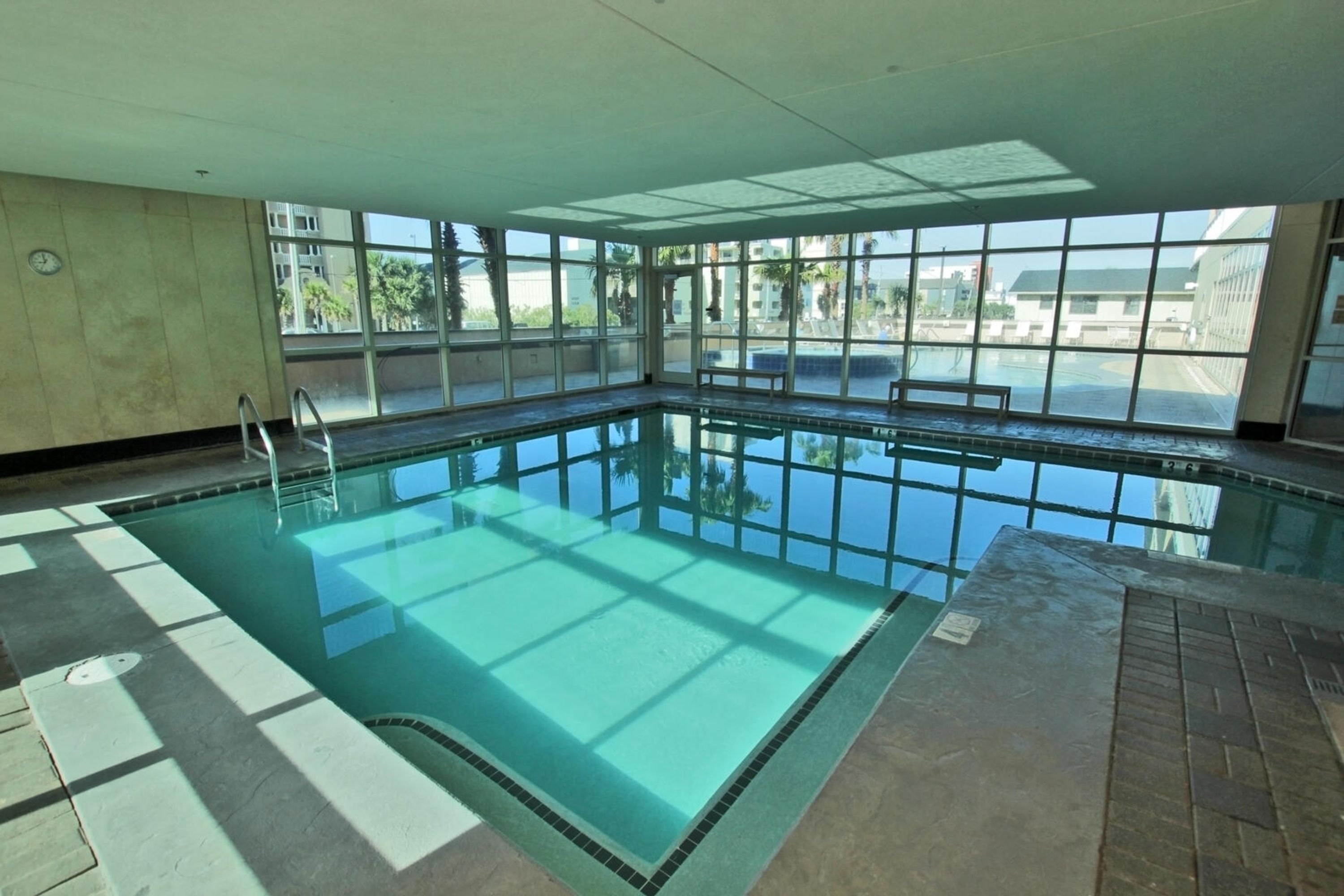 An indoor swimming pool with clear glass walls, a tiled floor, and a bench along the edge.