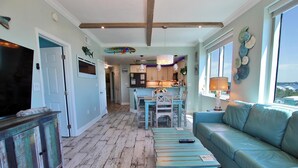 Amplia sala de estar con un sofá azul, una mesa de centro de madera y un televisor sobre un mueble. Se ve al fondo una cocina con armarios de madera y una zona de comedor con una mesa y sillas.