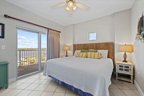 A bedroom with a large bed, two bedside lamps, a ceiling fan, a window with curtains, and a view of the outdoors.
