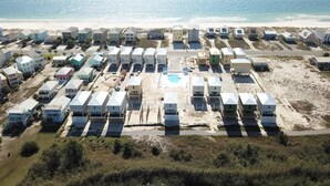 An aerial view of a coastal residential area with multiple houses, a swimming pool, and a beach in the background.